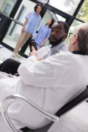 Photo for Senior specialist doctor discussing health care treatment with patient while showing pills bottle during consultation in hospital waiting area. Diverse people standing in lobby, medicine service - Royalty Free Image