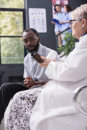 Photo for African american patient sitting on chair in hospital waiting area while medic explaining medication treatment during examination. Senior physician doctor showing pill bottles full with painkiller - Royalty Free Image