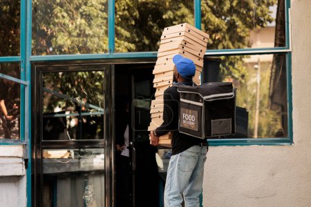 Téléchargez les photos : Courrier avec des boîtes à pizza pile debout devant l'immeuble de bureaux, livreur tenant ordre de la nourriture à l'extérieur. Service de livraison de déjeuner travailleur afro-américain livrant fastfood - en image libre de droit