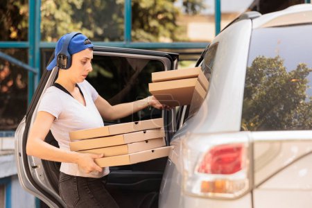 Photo for Woman delivering pizza to office by car side view, young fast food restaurant courier holding boxes pile. Pizzeria delivery service caucasian employee in headphones carrying lunch - Royalty Free Image