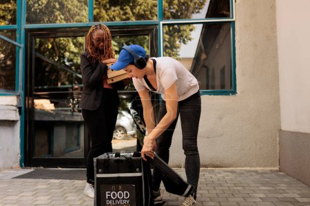 Photo for Courier in headphones taking out pizza boxes from thermal bag, customer receiving pizzeria order in front of office building outdoors. Food delivery service, woman delivering takeaway meal - Royalty Free Image
