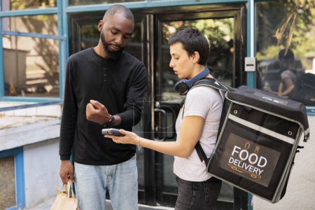 Photo for Client making contactless payment with smart watch for food delivery. Woman courier holding pos terminal device, customer paying for takeaway meal in front of office building medium shot - Royalty Free Image