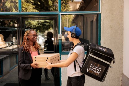 Téléchargez les photos : Travailleur de bureau afro-américain souriant recevant une commande de pizza à l'extérieur, femme prenant des paquets de repas empilés du courrier. Fille dans les écouteurs livraison repas à emporter, service de livraison de pizzeria - en image libre de droit