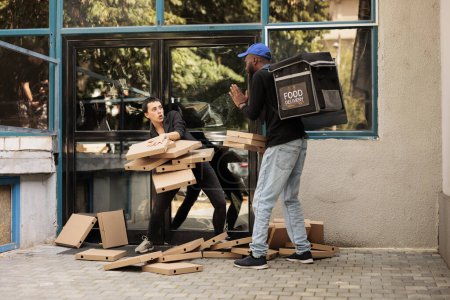 Téléchargez les photos : Femme perplexe attrapant la pile de boîtes à pizza tombant, courrier effrayé debout devant l'immeuble de bureaux. Homme afro-américain négligent livrant un repas à emporter au café, mauvais service de livraison - en image libre de droit