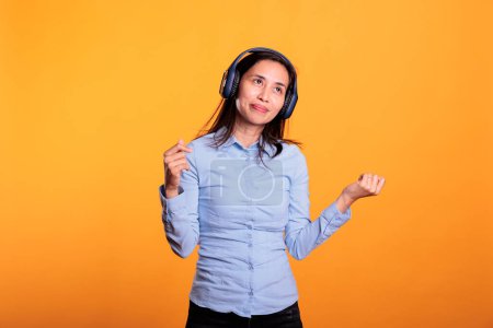 Téléchargez les photos : Femme asiatique confiante écoutant de la musique et dansant, profitant du temps de pause en studio sur fond jaune. Modèle souriant portant des écouteurs montrant des mouvements de danse drôles. Concept de divertissement - en image libre de droit