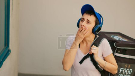 Photo for Tired food delivery employee yawning and ringing front door bell to deliver takeaway meal order from restaurant. Fastfood courier feeling sleepy and carrying backpack. Handheld shot. - Royalty Free Image