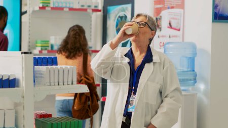 Photo for Health specialist drinking cup of coffee near pharmacy shelves, looking at supplements and pills in boxes. Checking packages leaflet of medicaments and vitamins, pharmaceutical products. - Royalty Free Image