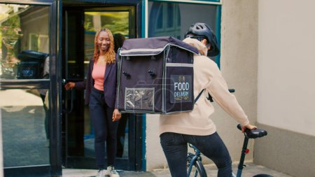 Foto de La persona de entrega rápida de comida da orden de comida en paquete a la mujer afroamericana, entregando comida de bolsa de papel en la puerta principal. Mensajera sosteniendo comida rápida para llevar almuerzo, servicio express. - Imagen libre de derechos