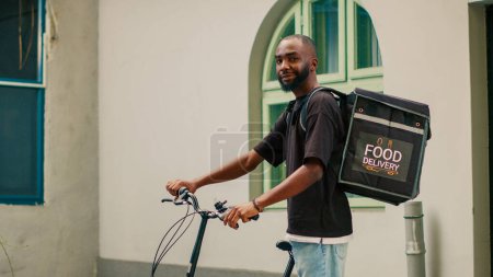 Photo for African american male courier riding bike to give meal package to customer, carrying backpack at front door. Deliveryman working with bicycle and delivering restaurant takeaway food. - Royalty Free Image