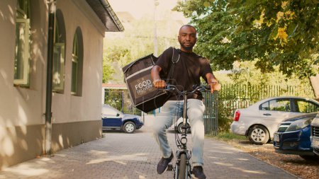 Photo for Male delivery courier riding bike with thermal backpack to give fastfood order package to clients. Going to front door entrance to deliver takeaway meal, restaurant delivery service. - Royalty Free Image