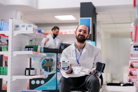 Téléchargez les photos : Portrait d'un pharmacien tenant un tonomètre médical prêt à mesurer l'hypertension des clients lors d'une visite de contrôle en pharmacie. Droguerie remplie de suppléments et de vitamines, service de médecine - en image libre de droit