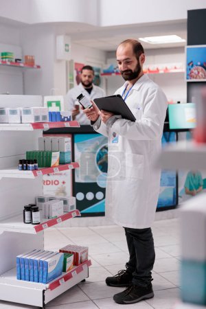 Foto de Farmacéutico mirando paquetes de pastillas que son responsables de llenar y etiquetar con precisión las recetas de medicamentos en el ordenador tableta en la farmacia. Servicio y concepto de apoyo a la salud - Imagen libre de derechos