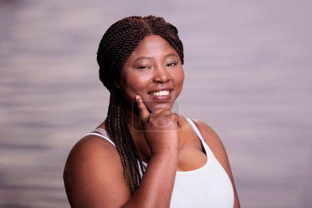 Foto de Afro americano sonriente belleza más modelo de tamaño apuntando a la piel limpia, sosteniendo el dedo en la mejilla y mirando a la cámara. Mujer atractiva posando en estudio con expresión facial positiva - Imagen libre de derechos