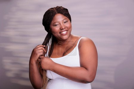 Photo for Body positive woman braiding long brown hair with ombre effect. Pretty curvy african american lady holding braids, looking at camera and expressing joyful emotions in studio - Royalty Free Image