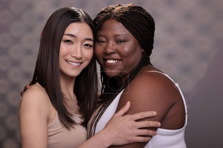 Téléchargez les photos : Deux femmes diverses embrassant démontrant la confiance, l'amour et l'autosoin. Belle afro-américaine et asiatique jeunes copines souriantes, embrassant ensemble et regardant la caméra - en image libre de droit