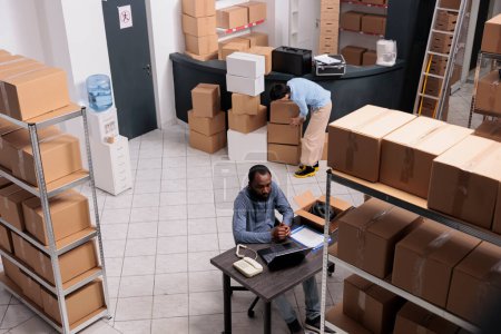 Photo for Top view of storehouse worker checking distribution logistics on laptop computer while preparing client package putting order in carton box. Employee shipping customer good, working in warehouse - Royalty Free Image