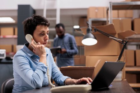 Photo for Storehouse manager talking at landline phone with remote supervisor discussing distribution logistics problem while looking at cargo stock on laptop. Employee working at order delivery in warehouse - Royalty Free Image