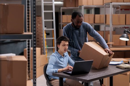 Photo for African american employee holding carton box while storehouse supervisor checking shipping client details before start delivering package. Diverse team working in delivery department in warehouse - Royalty Free Image