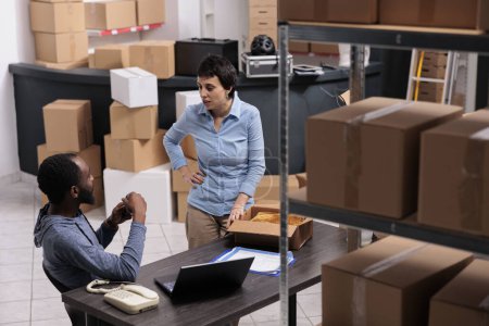 Photo for Warehouse supervisor preparing packages putting customer order in carton box while discussing shipping detalies with manager. Multi ethnic team working in storehouse delivery department - Royalty Free Image