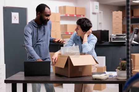 Photo for Storehouse workers discussing shipping logistics after putting client package in cardboard box after wrapping order in bubble wrap. Diverse employees working at merchandise transportation in warehouse - Royalty Free Image