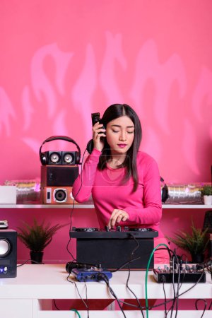 Photo for Asian artist with headphones having fun while playing techno music using professional mixer console, standing at dj table in studio with pink background. Musician performing electronic song in club - Royalty Free Image