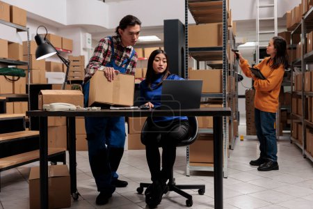 Photo for Sotrehouse employees doing delivery route optimization on laptop. Warehouse asian man and woman coworkers preparing parcel for shipment and checking orders checklist on computer - Royalty Free Image