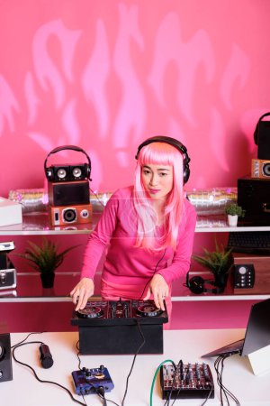 Téléchargez les photos : Artiste aux cheveux roses portant un casque tout en jouant de la musique électronique à l'aide d'une console de mixage professionnelle, appréciant jouer dans une boîte de nuit. Musicien asiatique debout à dj table mélange sons - en image libre de droit