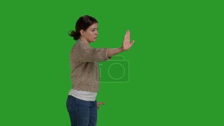 Photo for Side view of young woman raising palm as rejection gesture, expressing disapproval and disagreement symbol. Female model showing stop talking sign with hand, refuse and reject on greenscreen. - Royalty Free Image