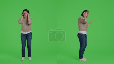 Photo for Happy woman listening to music on headset, having fun in studio over green screen backdrop. Young person enjoying sound and mp3 song on camera, using wireless headphones on full body background. - Royalty Free Image