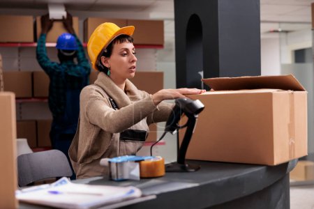 Photo for Retail storehouse manager packing cardboard box at counter desk and closing package. Caucasian warehouse worker wearing safety helmet preparing customer parcel for delivery - Royalty Free Image
