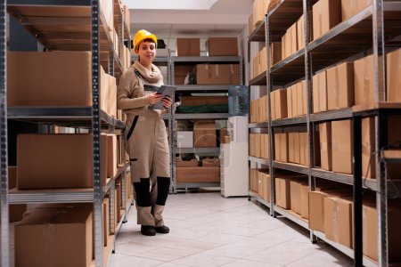 Photo for Delivery service storehouse worker checking cardboard boxes on shelf and looking at camera. Smiling woman in logistic storage protective uniform checking operations on digital tablet - Royalty Free Image