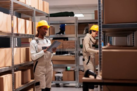 Photo for Warehouse manager supervising freight inventory and checking audit application. African american woman standing in storehouse near cardboard boxes shelf and using digital tablet - Royalty Free Image