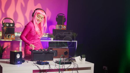 Téléchargez les photos : Asian musician with headphones playing electronic song at mixer console, standing at dj table having fun with fans at night in club. Artist performing techno music in studio over pink background - en image libre de droit