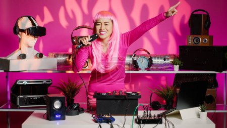 Téléchargez les photos : Cheerful performer having fun mixing techno sounds in night club, playing music using professional turntables. Asian artist standing at dj table dancing while celebrating techno album with fans - en image libre de droit