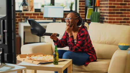 Photo for Smiling girl preparing to eat slice of pizza at tv, sitting on couch and watching comedy movie. Cheerful modern woman eating dinner from fast food meal delivery, drinking alcohol at home. - Royalty Free Image