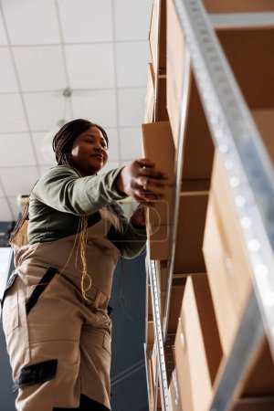 Photo for Stockroom employee taking out cardboard box from metallic shelf, preparing clients orders in storage room. Warehouse worker wearing industrial overall working at goods delivery - Royalty Free Image