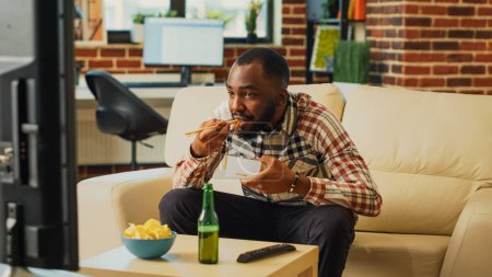 Foto de Happy guy eating noodles using chopsticks at home, opening bottle of beer and feeling relaxed at television. Smiling adult enjoying asian delivery meal for dinner, watching show on tv. - Imagen libre de derechos