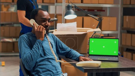 Téléchargez les photos : Homme entrepreneur parlant sur téléphone fixe et en utilisant greenscreen sur PC, travaillant avec écran chromatique isolé à l'entrepôt. Storehouse propriétaire avec téléphone et copyspace vierge. - en image libre de droit