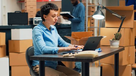 Photo for Female owner doing quality control before shipping goods in cardboard boxes, taking merchandise from warehouse shelves. Woman checking stock and doing distribution work. Handheld shot. - Royalty Free Image