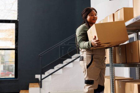 Photo for Supervisor standing on ladder holding cardboard box, working at products delivery in warehouse. Storage room employee preparing customers orders, checking packages in storehouse - Royalty Free Image