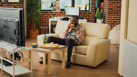 Foto de Cheerful adult taking bite of pizza at home, eating takeaway food delivery and drinking alcohol. Young relaxed man having fun watching action movie on television, eating snacks. - Imagen libre de derechos