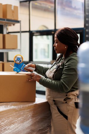 Photo for African american manager preparing clients packages for delivery using adhesive tape, doing products quality control in storehouse. Stockroom employee working at online orders in storage room - Royalty Free Image