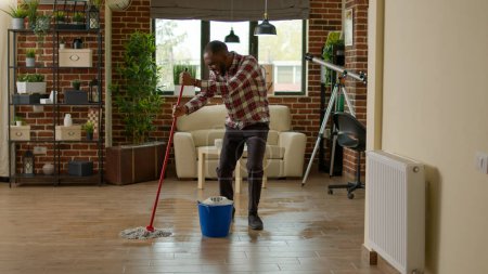Téléchargez les photos : Cheerful boyfriend dancing and washing floors with all purpose cleaner, using mop to sweep dust and dirt in living room. Young man using cleaning tool to do housework and chores. - en image libre de droit