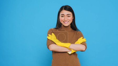 Téléchargez les photos : Femme de ménage souriante portant des gants en caoutchouc jaune debout avec le bras croisé en studio sur fond bleu. gai asiatique housekepper pris grand soin dans client logement devoirs - en image libre de droit