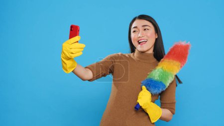 Téléchargez les photos : Smiling housekeeper holding colorful feather dust while taking selfie with mobile phone, standing in studio over blue background. Cheerful maid using high-quality detergents and rubber gloves - en image libre de droit