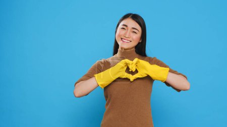 Téléchargez les photos : Happy asian maid wearing yellow gloves while doing heart shape in front of camera after finishing cleaning houses for customers. Housekeeper primary responsibilities is to maintain cleanliness - en image libre de droit