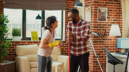 Photo for Life partners doing cleaning chores together in apartment, mopping floors and sweeping dust off. Diverse people enjoying spring cleaning session with mop and furniture polish. Handheld shot. - Royalty Free Image