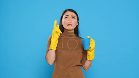 Foto de Pensive thoughtful maid holding cup of coffee standing over blue background in studio, thinking at solution to solve cleaning problem. Housekepper providing top-notch cleaning services to her clients. - Imagen libre de derechos