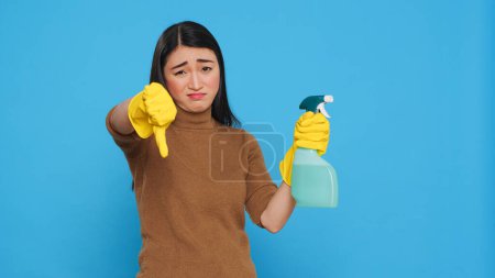 Téléchargez les photos : Upset unhappy housekeeper holding hygiene detergent spray doing disapproval sign studio. Maid is always professional in her cleaning while showing thumbs down gesture over blue background - en image libre de droit