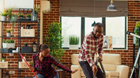 Foto de Happy husband and wife enjoying cleaning with mop and vacuum cleaner, singing and listening to music at home. Young partners mopping and vacuuming tiles in apartment. Tripod shot. - Imagen libre de derechos